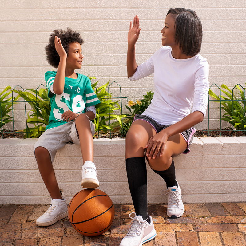 Dziecko i kobieta przybijają sobie piątkę, siedząc po meczu koszykówki; kobieta ma na sobie podkolanówki (skarpety) uciskowe. Dziecko i kobieta przybijają sobie piątkę, siedząc po meczu koszykówki; kobieta ma na sobie podkolanówki (skarpety) uciskowe.